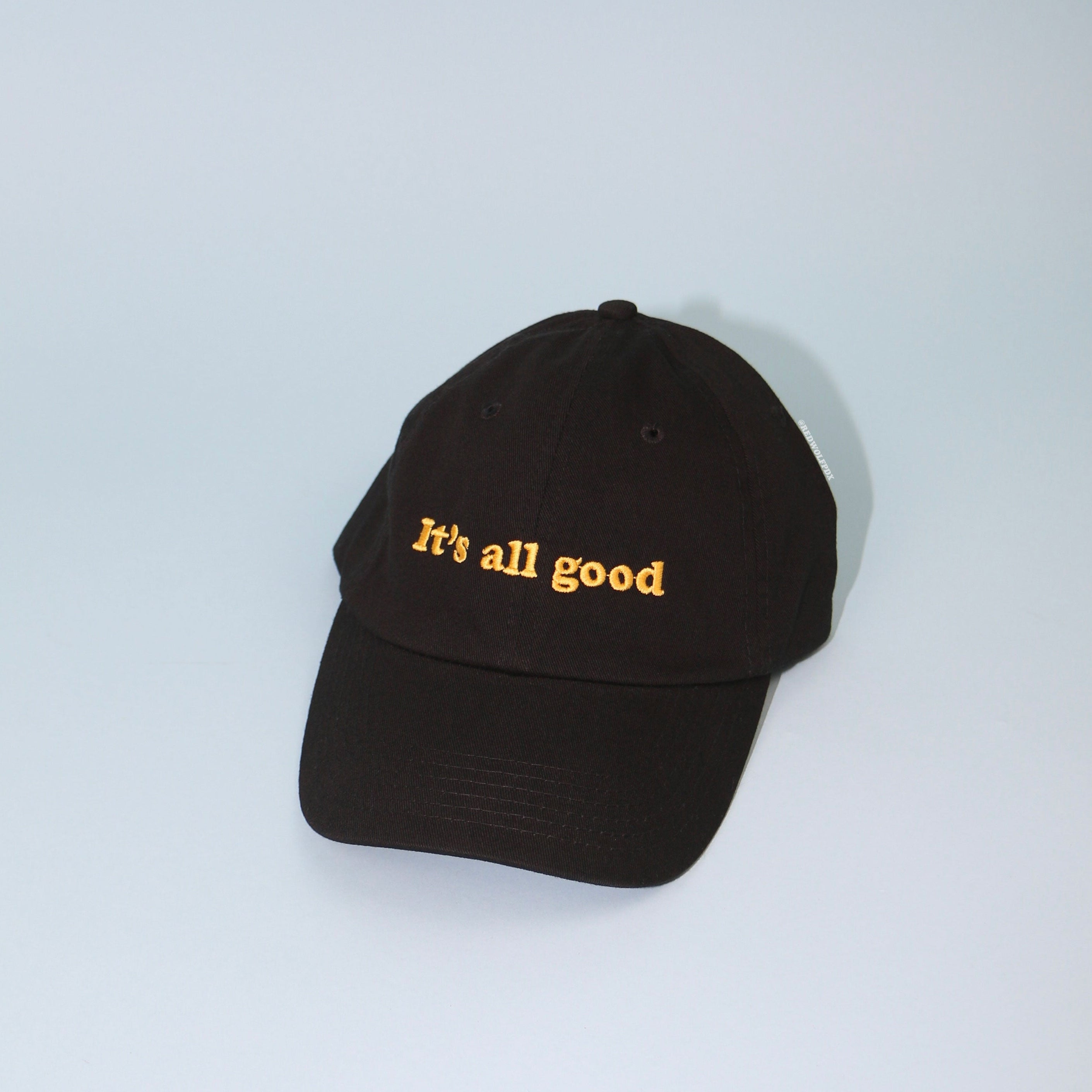 Black baseball cap embroidered with the text 'It's all good' in yellow thread on blue backdrop