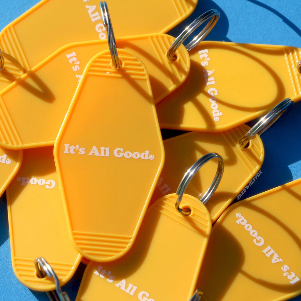 Pile of yellow retro motel style keychains with the text 'It's All Good' with smiley face graphic in white ink printed across it against a blue background
