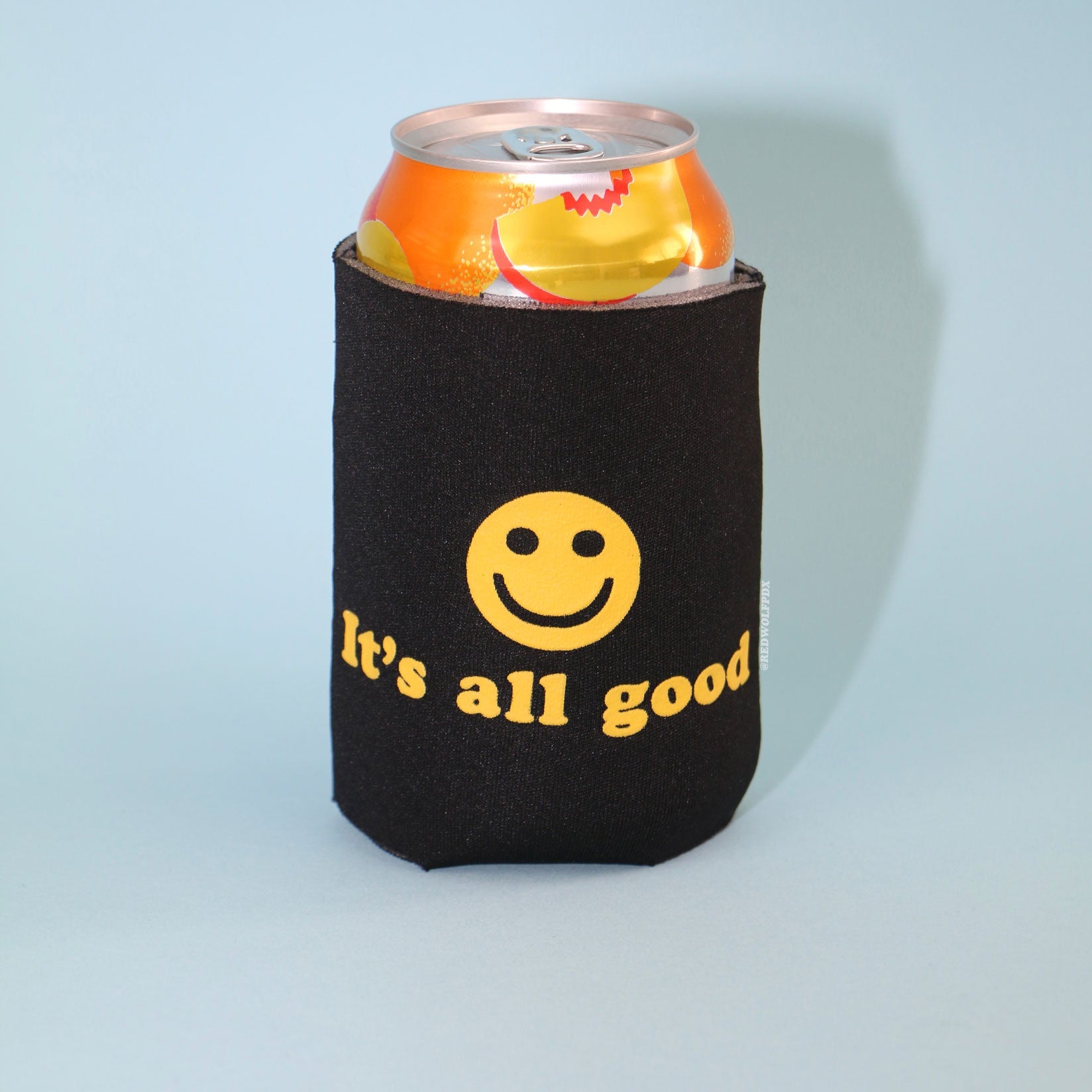 12 oz can beverage in a black neoprene drink sleeve with text 'It's all good' and smiley face graphic in yellow ink screen printed across it on a blue backdrop