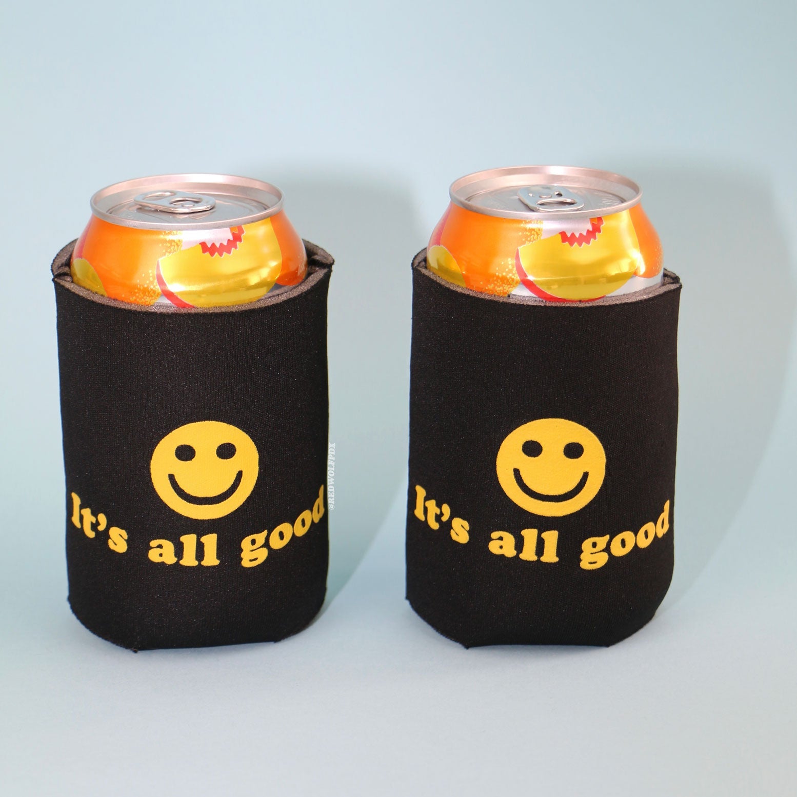 two black neoprene drink sleeves with text 'It's all good' with smiley face screen printed in yellow ink across both holding 12 oz. can beverages on a light blue backdrop.