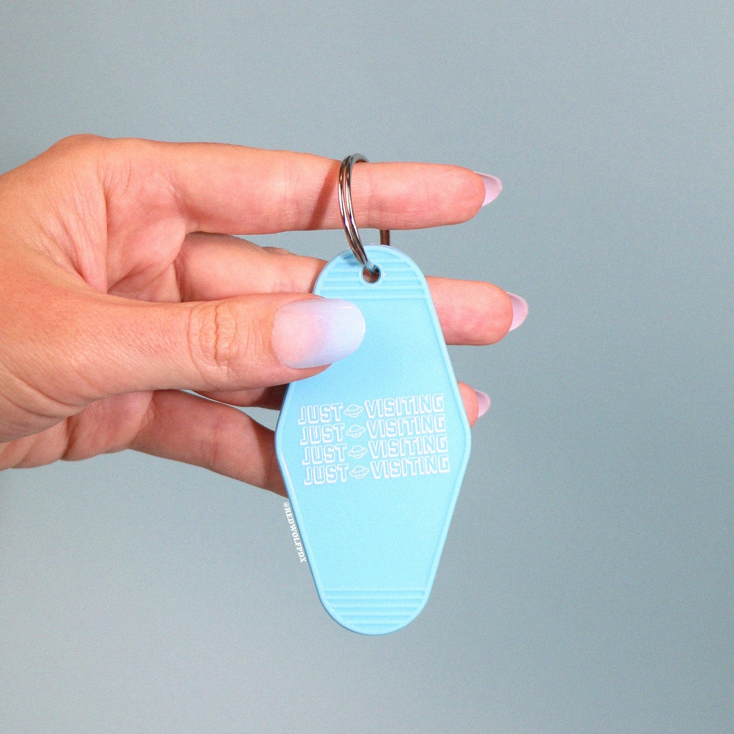 baby blue retro motel keychain with text 'JUST VISITING' in white ink printed across being held against blue backdrop