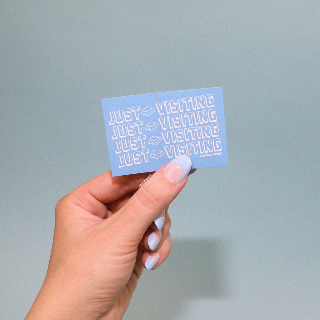 retro baby blue rectangle sticker with text 'JUST VISITING' being held in hand against a blue backdrop