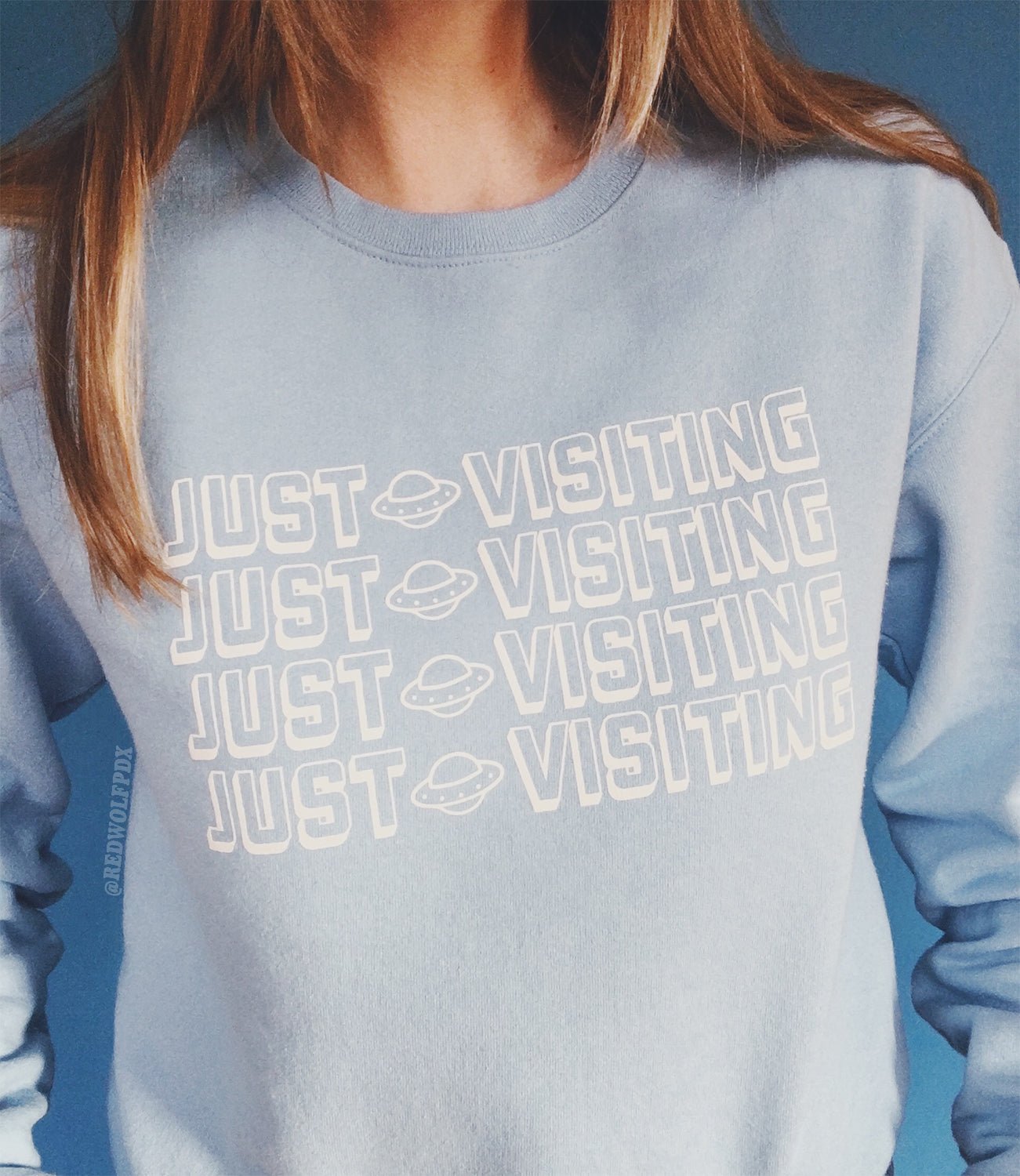 Retro baby blue crewneck sweatshirt with text 'JUST VISITING' across front of chest being worn against  blue backdrop