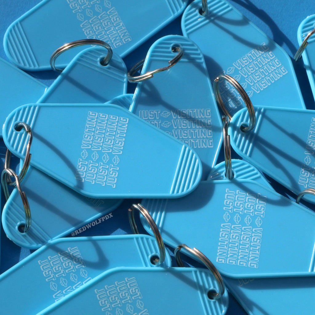 A pile of baby blue retro motel style keychains with the text 'JUST VISITING' printed across them
