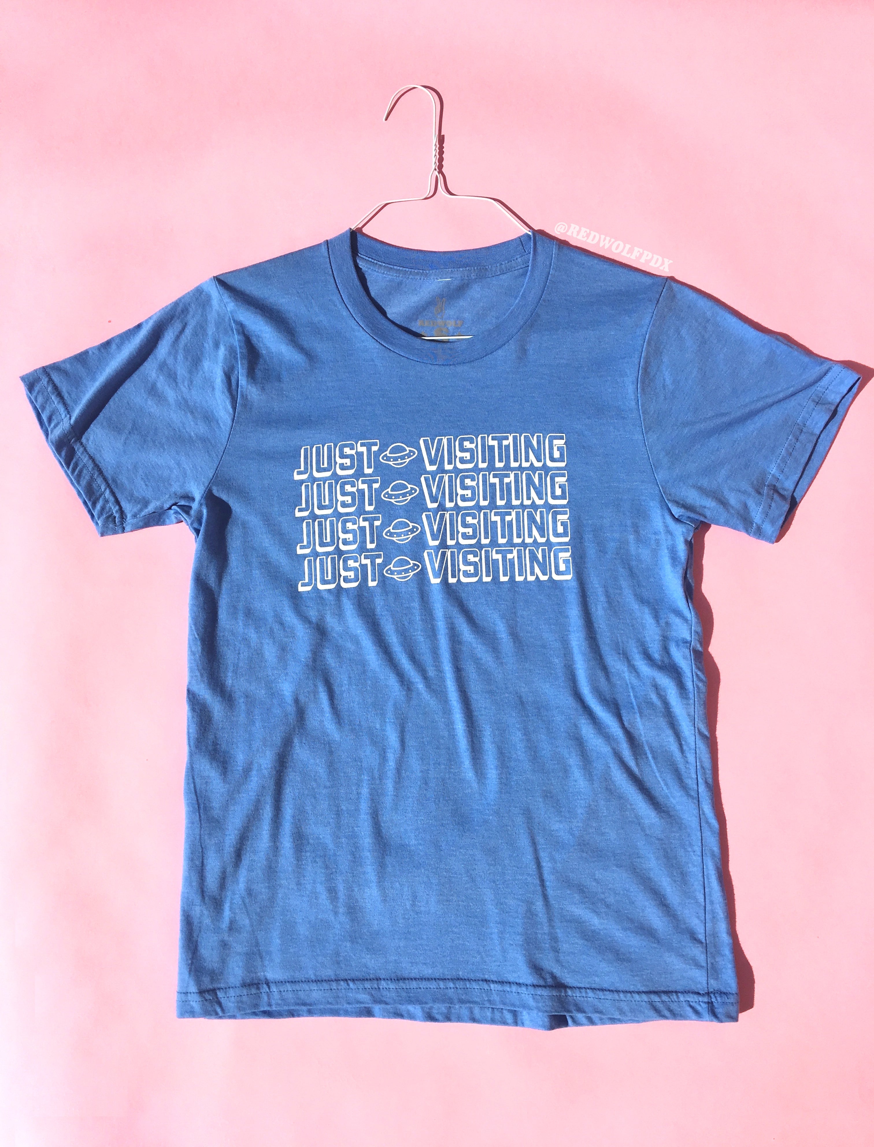 baby blue graphic tee with text 'JUST VISITING' with UFO graphic in white ink screen printed down the front against a light pink background