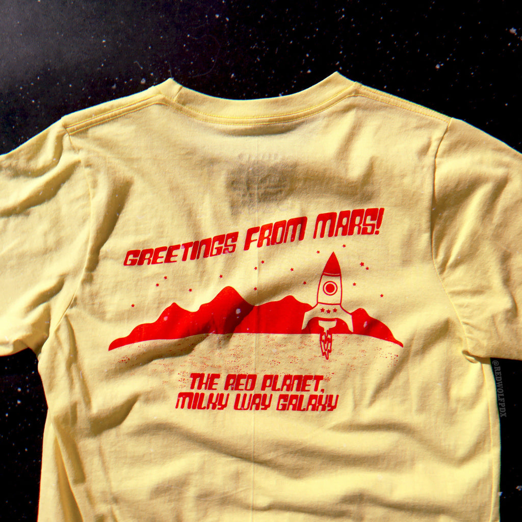 Powder yellow graphic tee with text 'Greetings from Mars!' with space illustration across the back in a vintage orange red ink against a space background