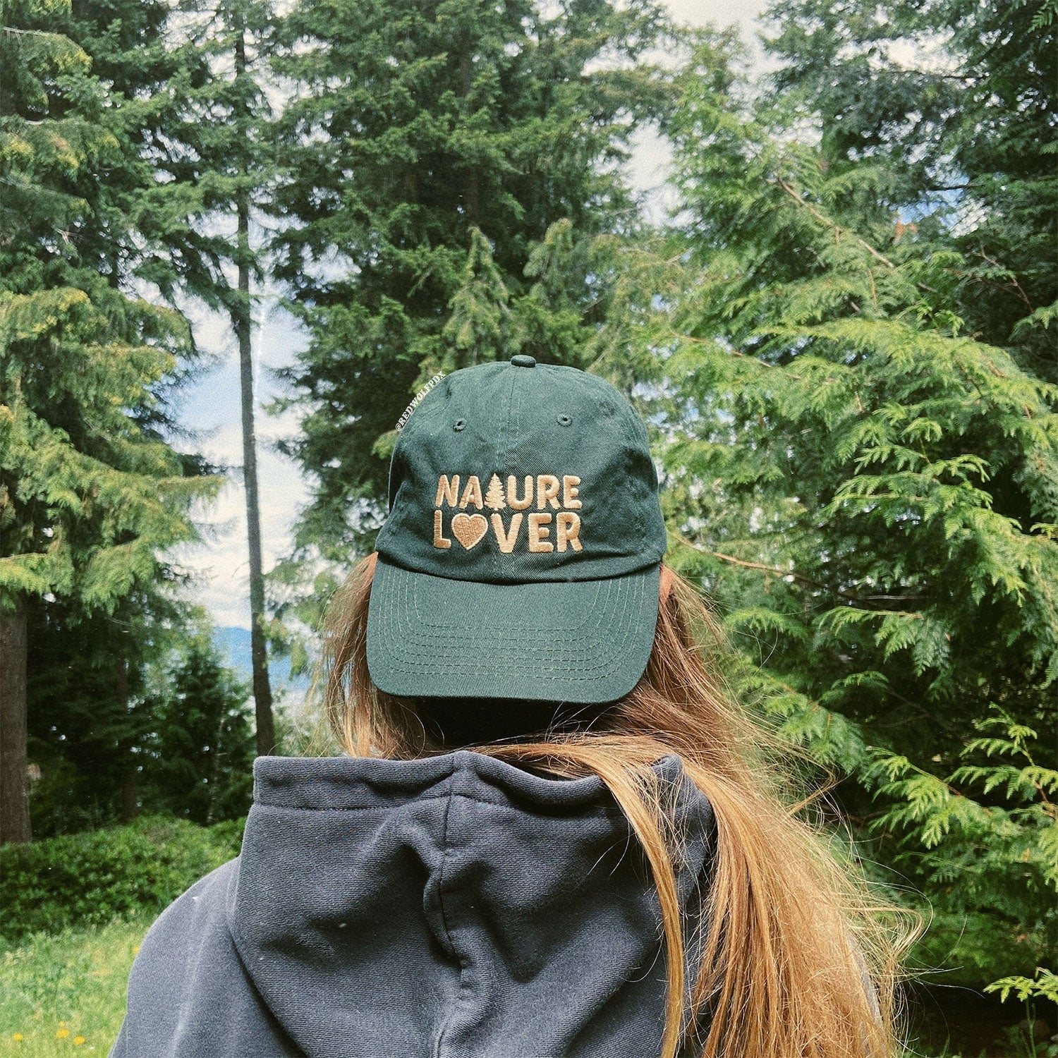 forest green baseball cap with text 'NATURE LOVER' embroidered with beige thread across the front being worn backwards against a nature landscape