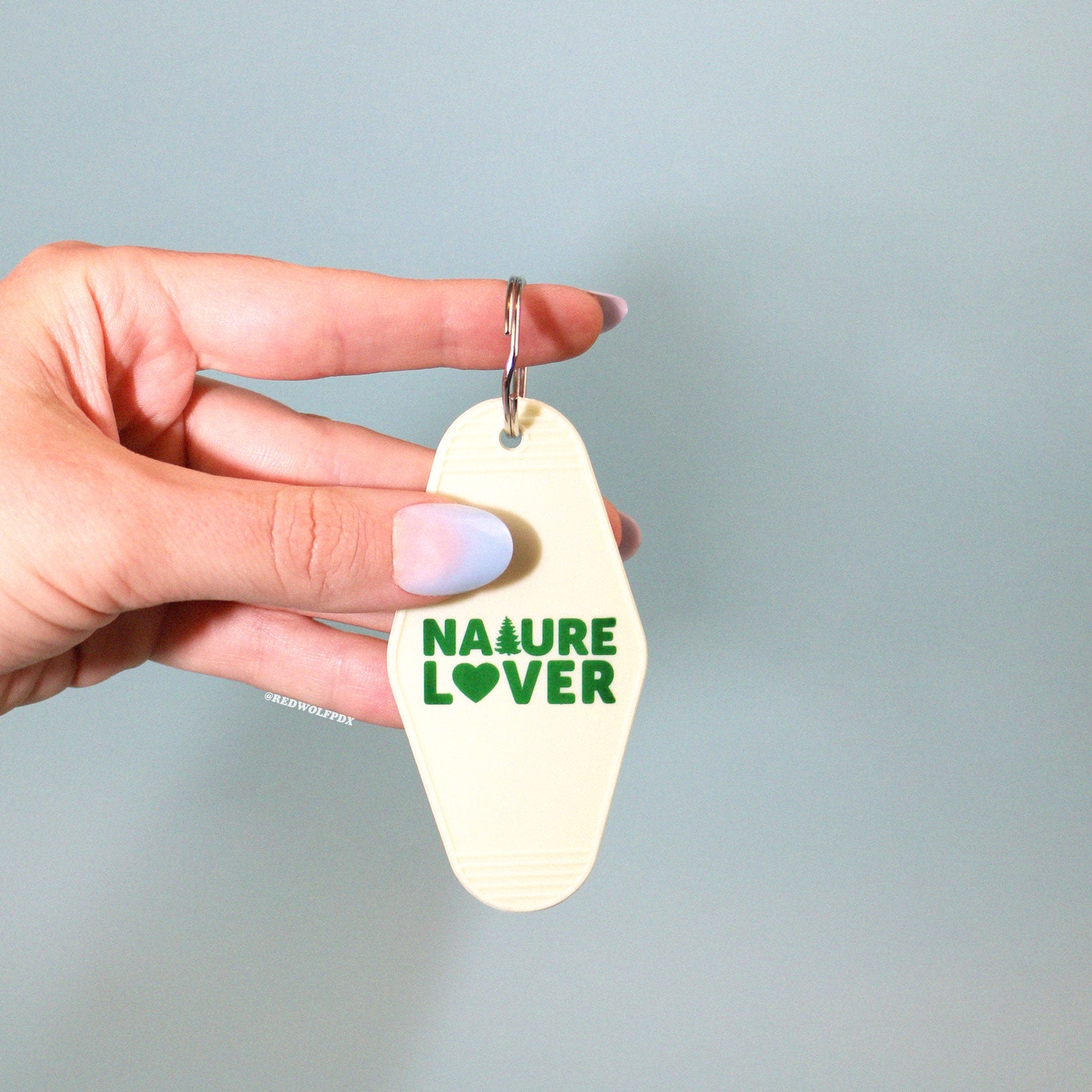 A vintage white keychain with the text 'NATURE LOVER' in dark green, shaped like a motel key being held against a blue backdrop