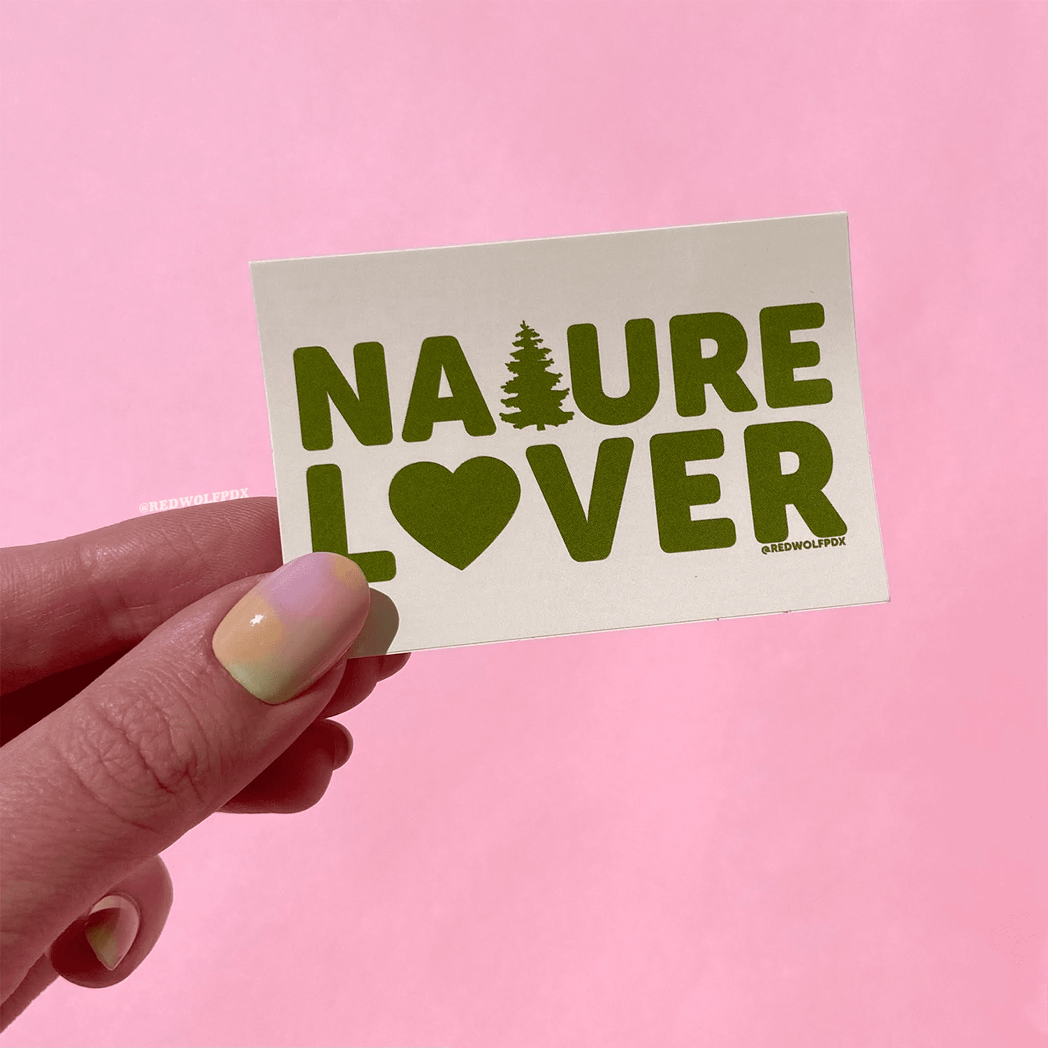 Rectangle beige sticker with text 'NATURE LOVER' in green ink being held against a pink backdrop