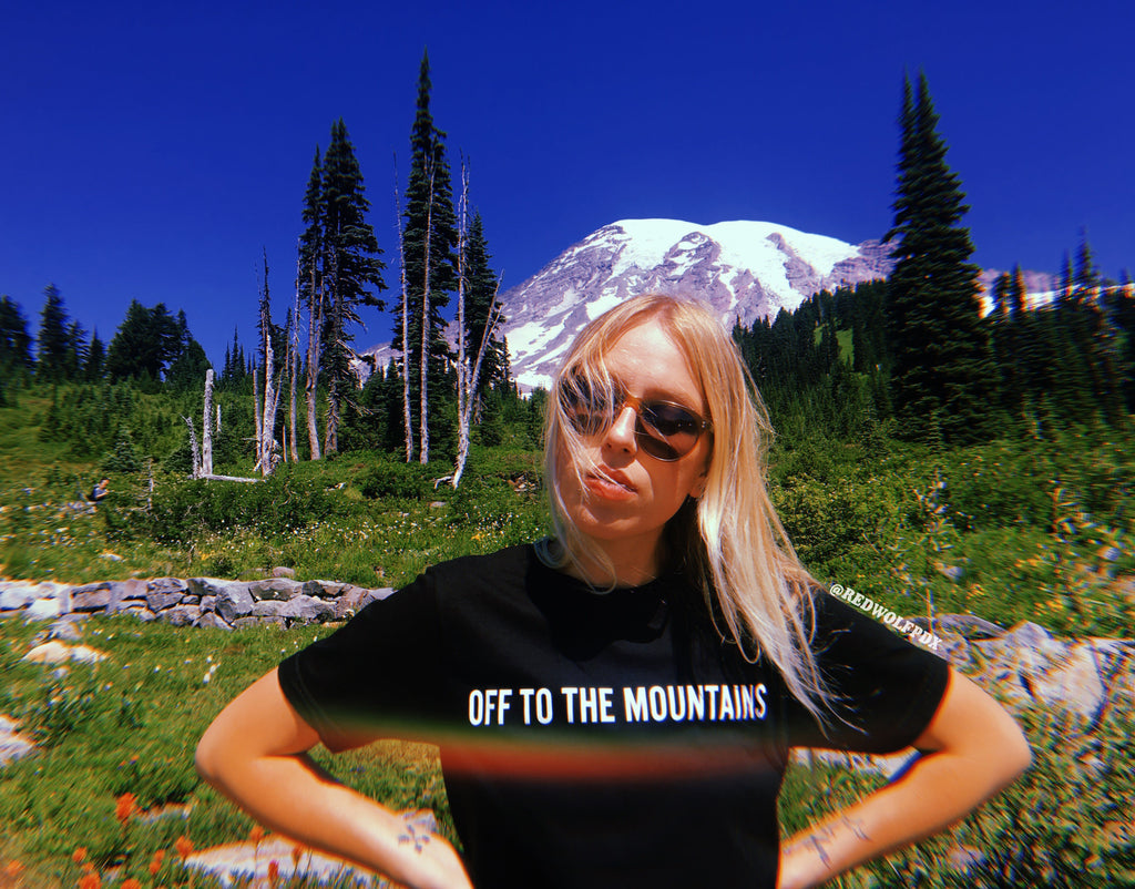 black graphic tee with text 'OFF TO THE MOUNTAINS' being worn with mount rainier and wildflowers in background