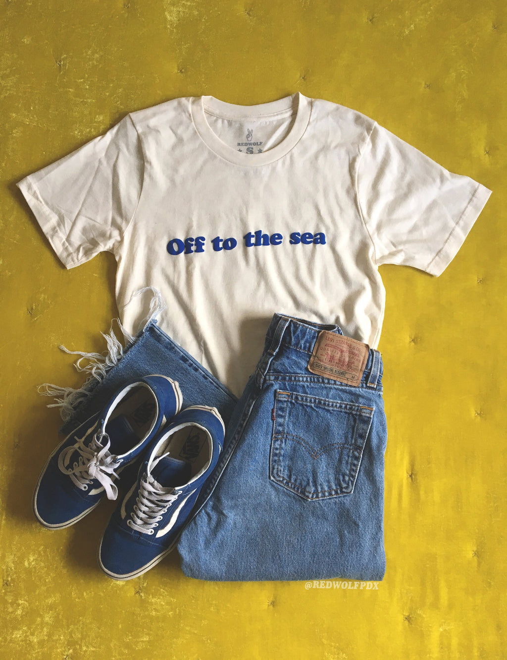Beige white graphic tee with text 'Off to the sea' in blue ink screen printed across in flat lay with blue jeans and sneakers on a vintage mustard yellow velvet blanket