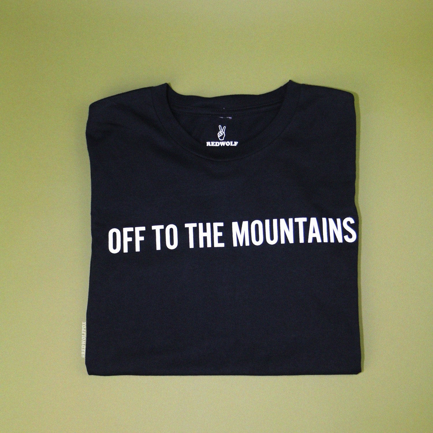 off to the mountains graphic tee folded on green backdrop