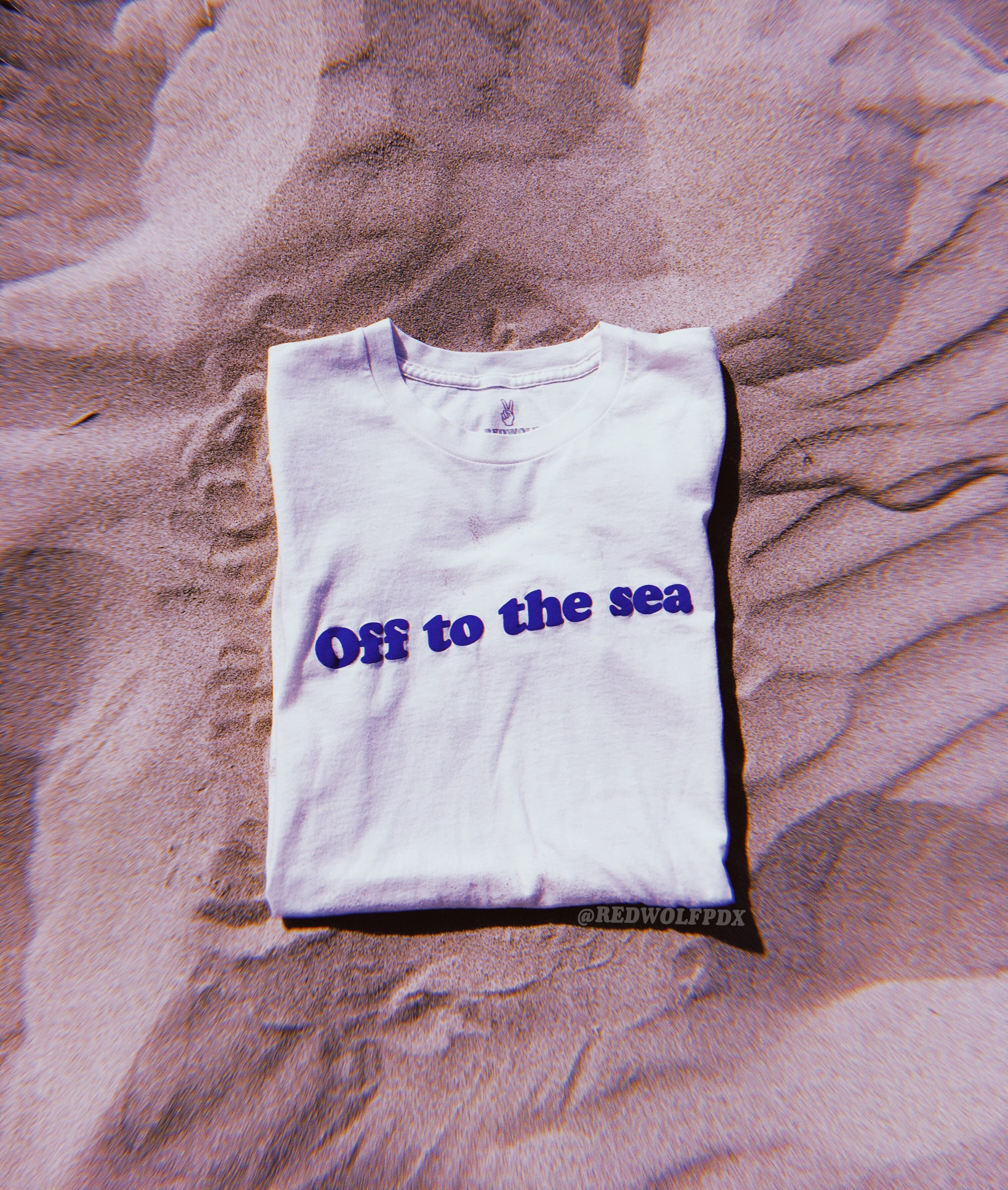 Folded beige white graphic tee with text 'Off to the sea' screen printed with raised blue ink across the front laying on sand 