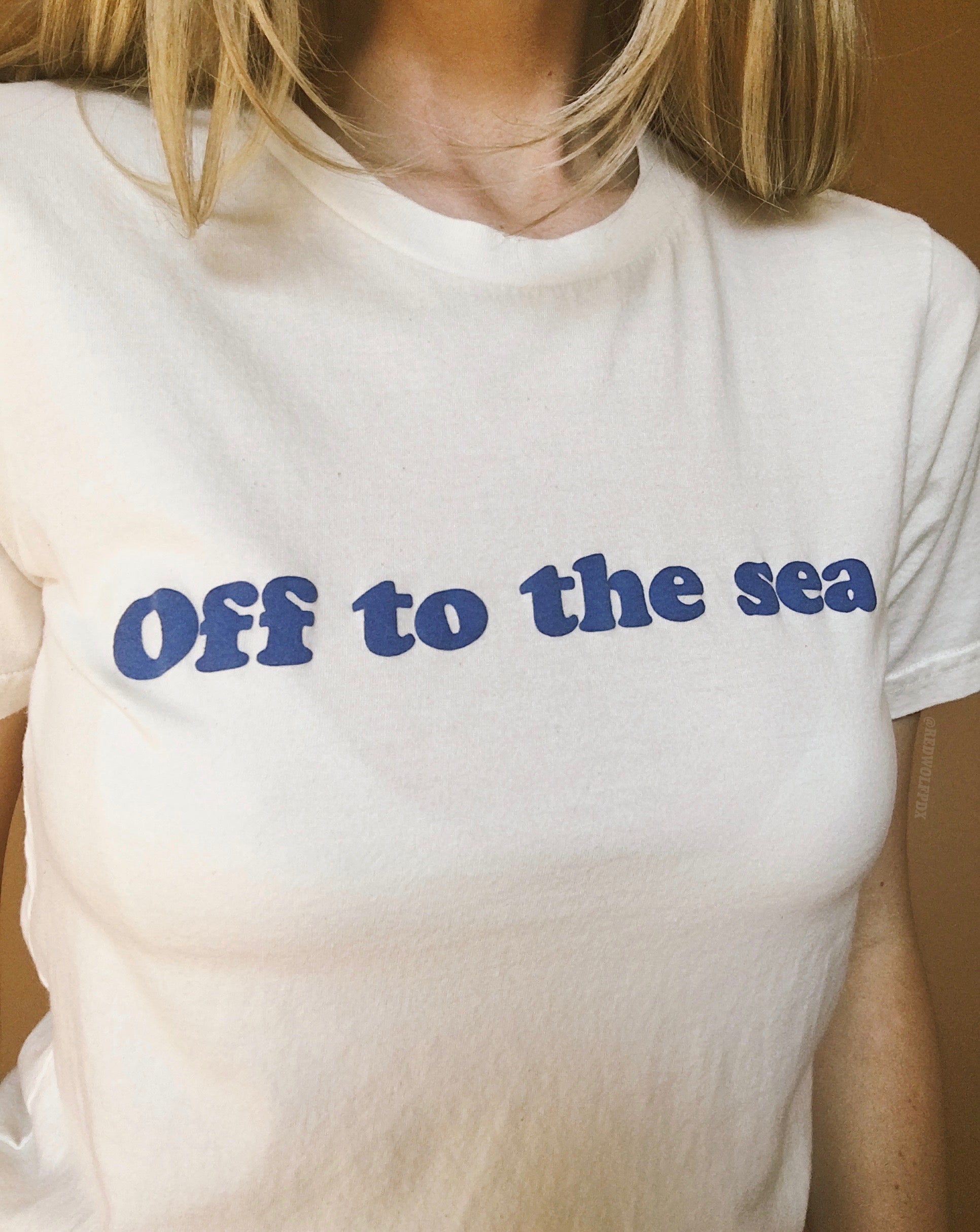 Beige white graphic tee with text 'Off to the sea' screen printed with raised blue ink across the front with a tan brown backdrop