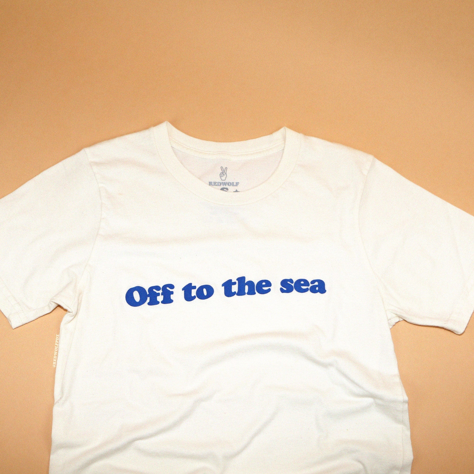 Beige white graphic tee with text 'Off to the sea' screen printed with raised blue ink across front on a beige tan background