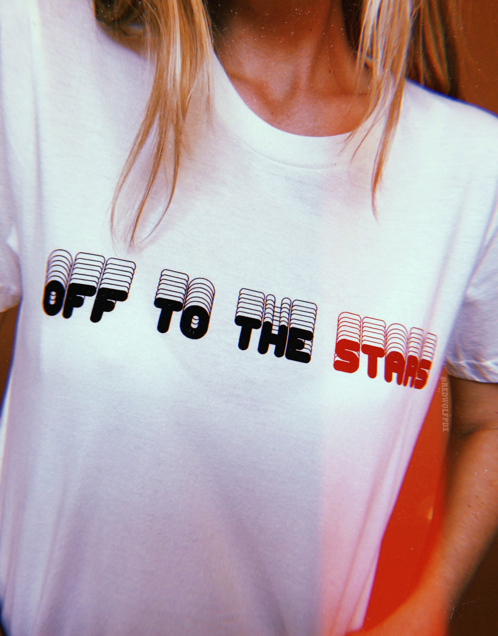 white tee with text 'OFF TO THE STARS' screen printed across the chest in black ink being worn against a brown backdrop