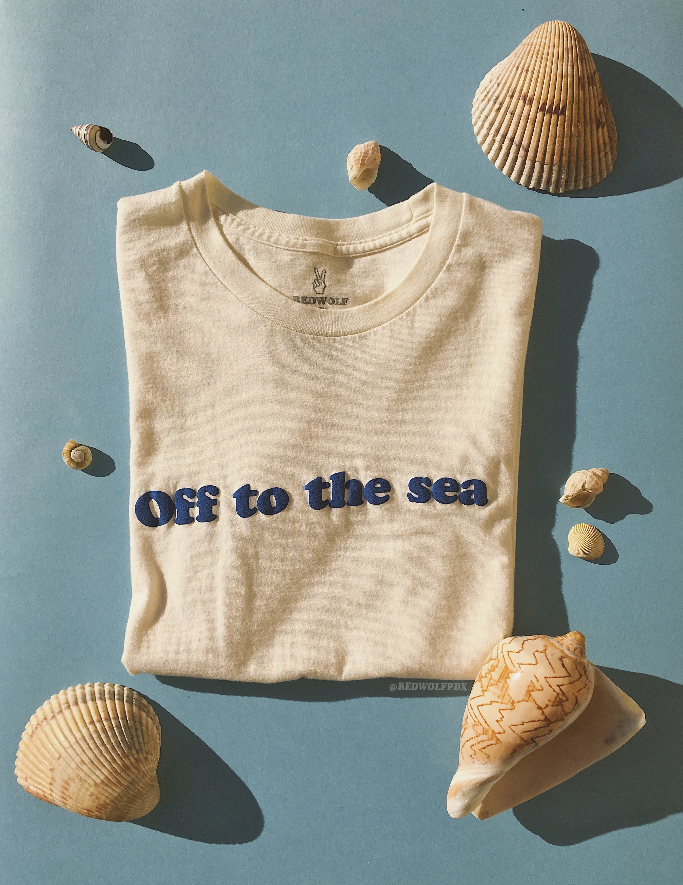 Folded beige white graphic tee with text 'Off to the sea' in raised blue screen printed ink across chest on a blue backdrop surrounded by seashells