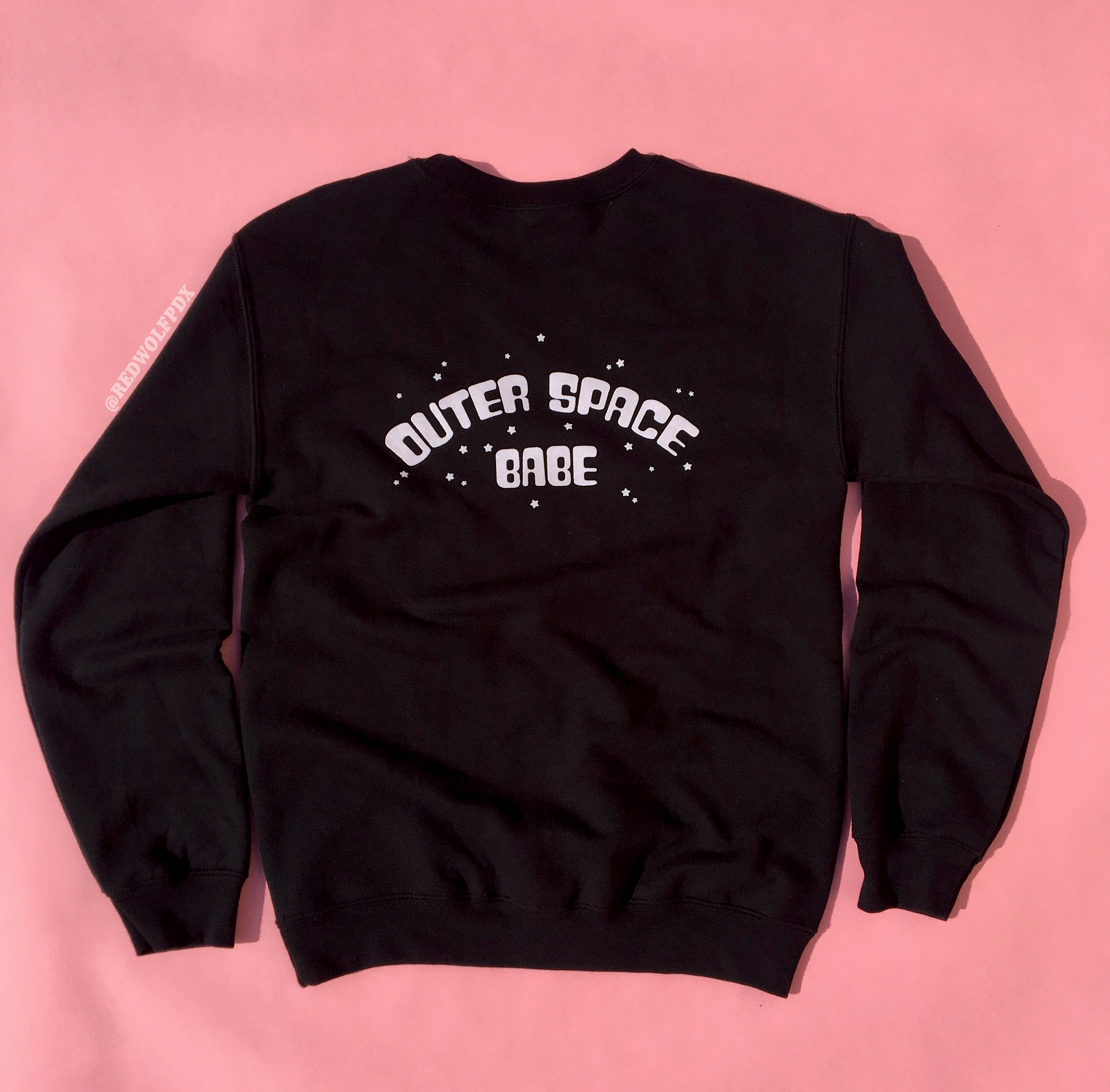 black crewneck sweatshirt with text 'OUTER SPACE BABE' in light pink ink with little stars illustration graphic screen printed across the back against a pink backdrop