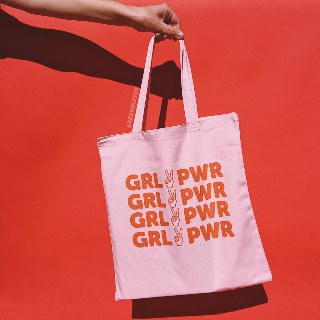 light pink canvas tote bag with text 'GRL PWR' in red orange ink screen printed down the front being held against a red backdrop