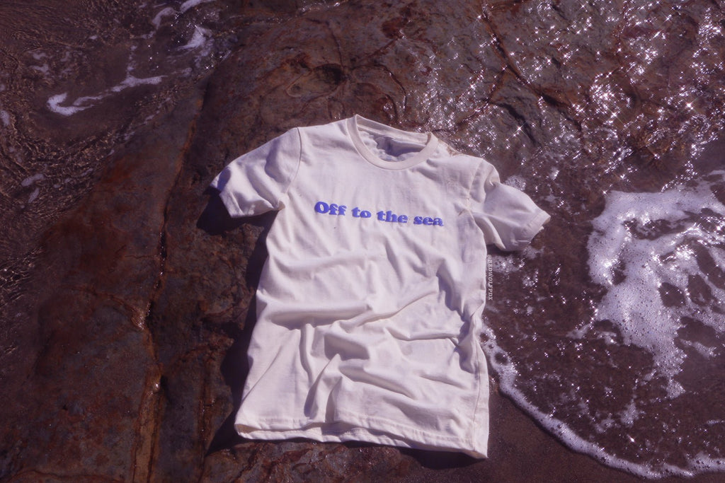 Beige white graphic tee with text 'Off to the sea' screen printed with raised blue ink across the front on a a beach