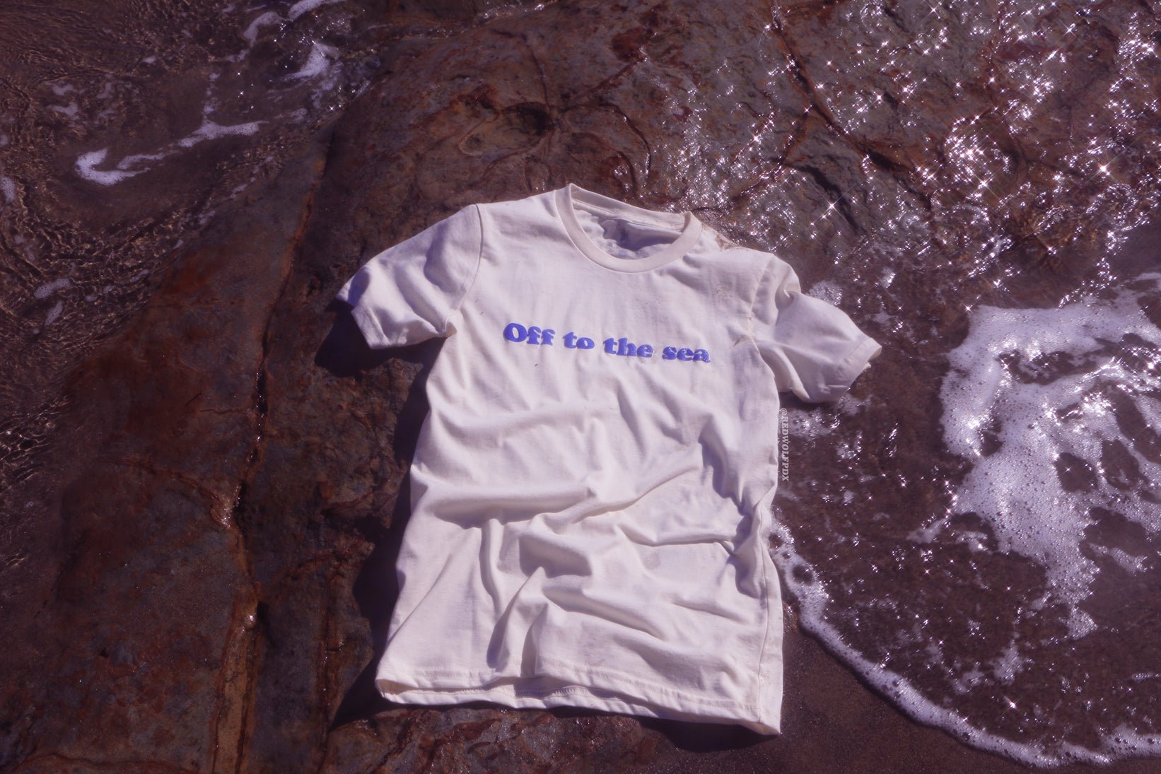 Beige white graphic tee with text 'Off to the sea' screen printed with raised blue ink across the front on a a beach