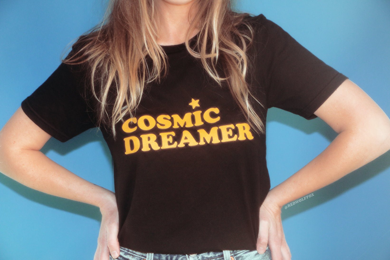 black graphic tee with text 'COSMIC DREAMER' in yellow ink screen printed across chest being worn against blue backdrop