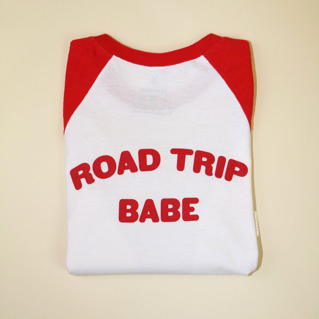 Folded red and white raglan tee with text 'ROAD TRIP BABE' in red ink screen printed across the back on a beige backdrop