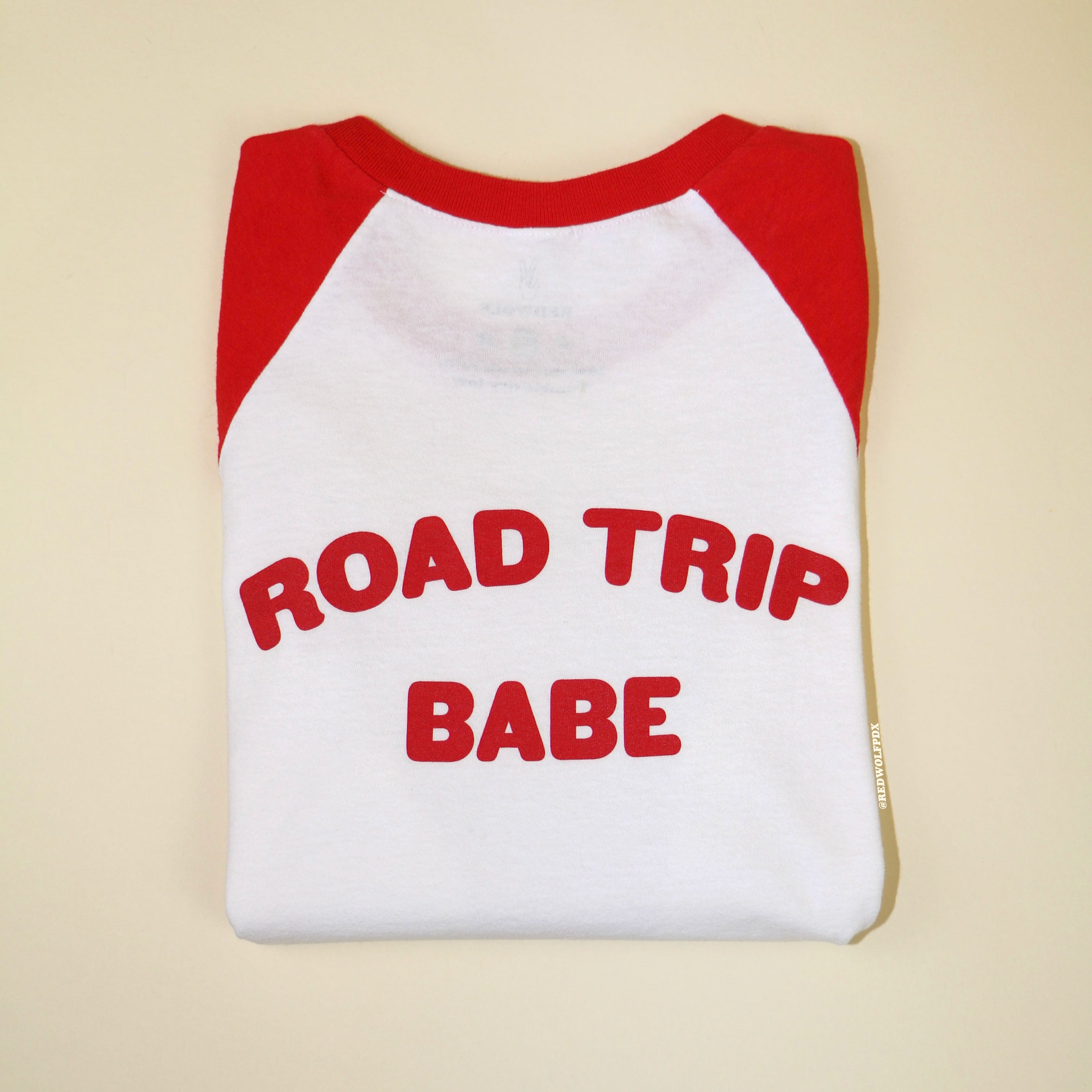 Folded red and white raglan tee with text 'ROAD TRIP BABE' in red ink screen printed across the back on a beige backdrop