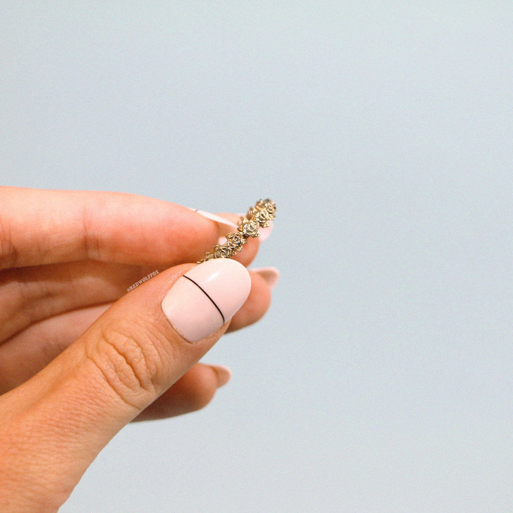 A close-up image of a hand holding a rose wreath ring with a brass finish, on a light background.