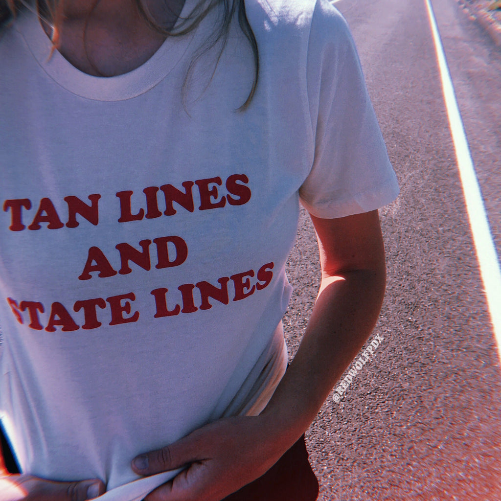 white tee with text 'TAN LINES AND STATE LINES' screen printed in red ink across chest being worn on side of road