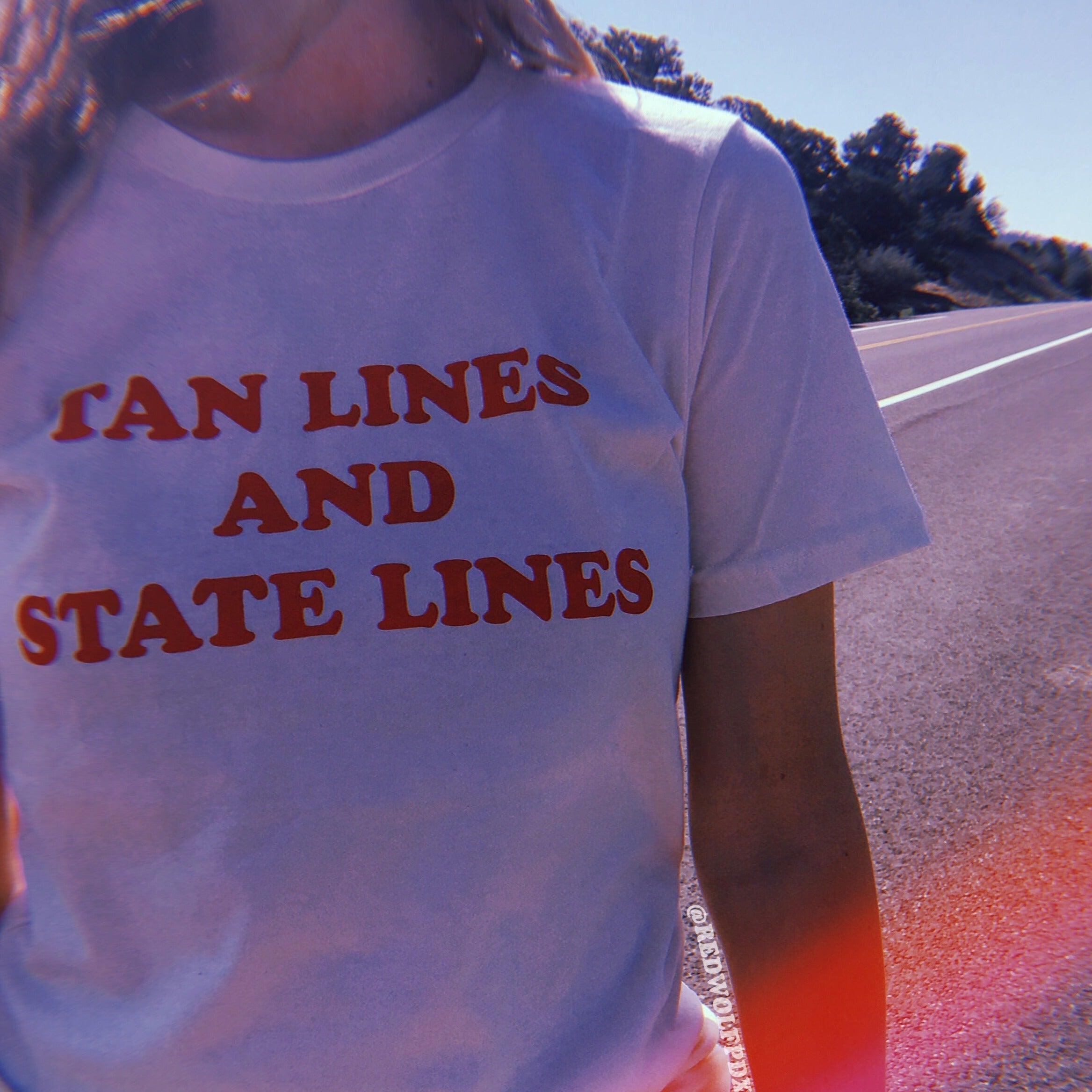 'TAN LINES AND STATE LINES' text graphic on white tee being worn against backdrop of road at national park