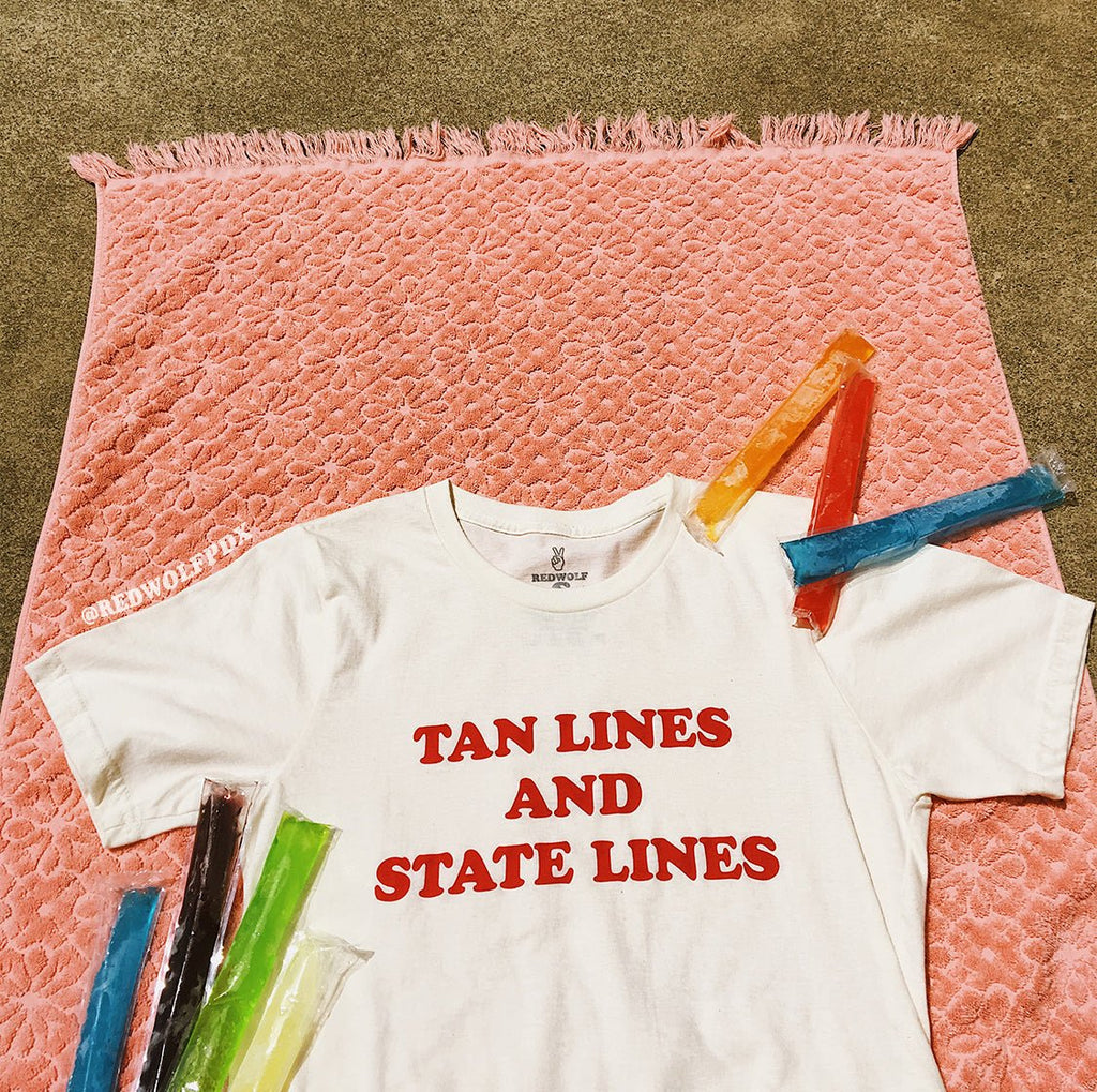 white tee with text 'TAN LINES AND STATE LINES' in red ink screen printed across chest on a vintage pink beach towel with colorful frozen pops