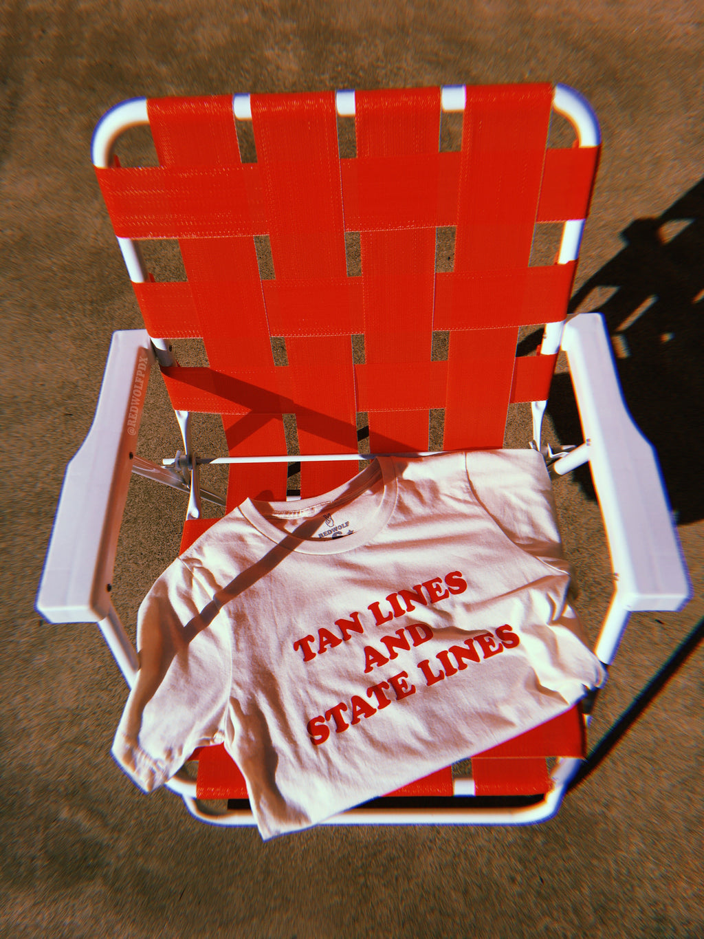Folded beige graphic tee with text 'TAN LINES AND STATE LINES' in orangey red screen printed on chest rested on red lawn chair.