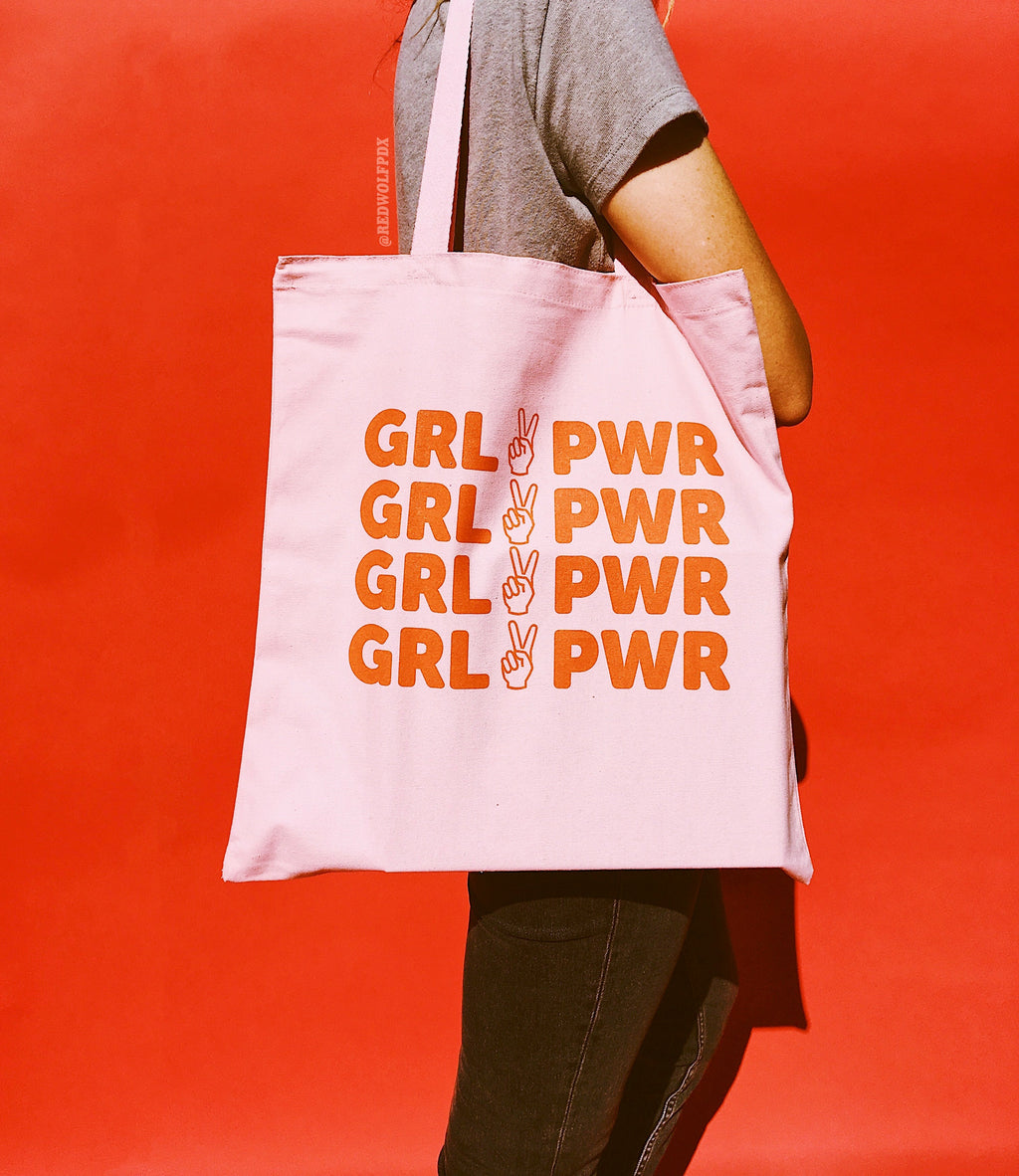 light pink canvas tote bag with text 'GRL PWR' in red orange ink screen printed across front being worn against a red backdrop