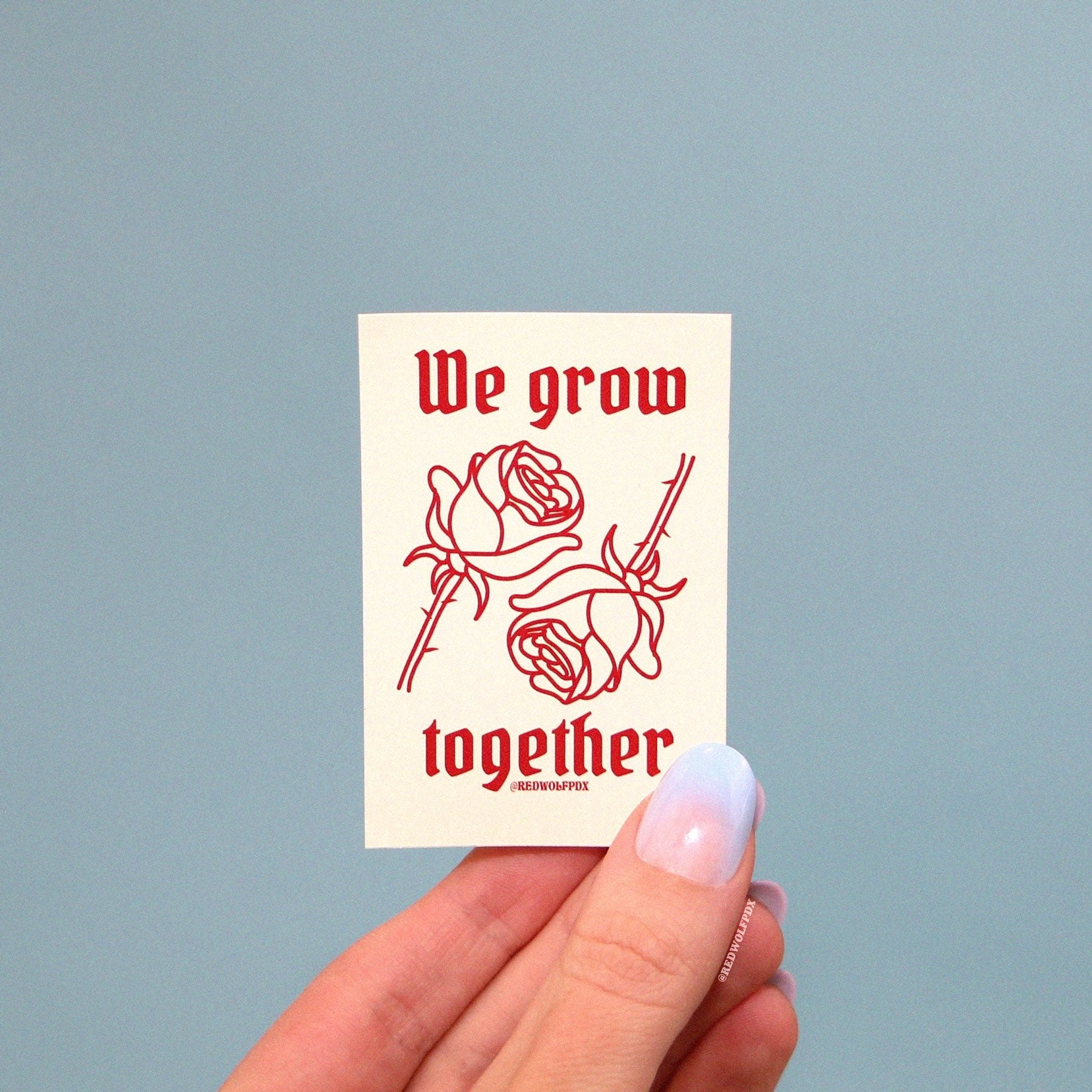 we grow together sticker with rose illustration being held up to a baby blue backdrop