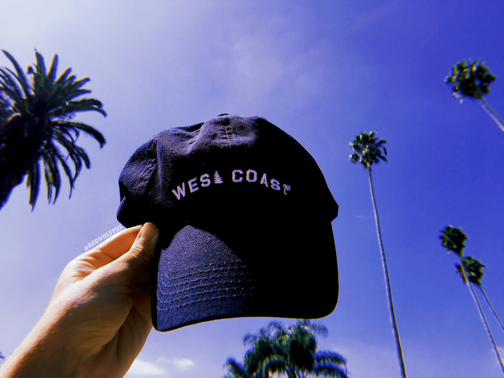 navy blue baseball cap with text 'WEST COAST' embroidered with white thread being held up to blue sky and palm trees