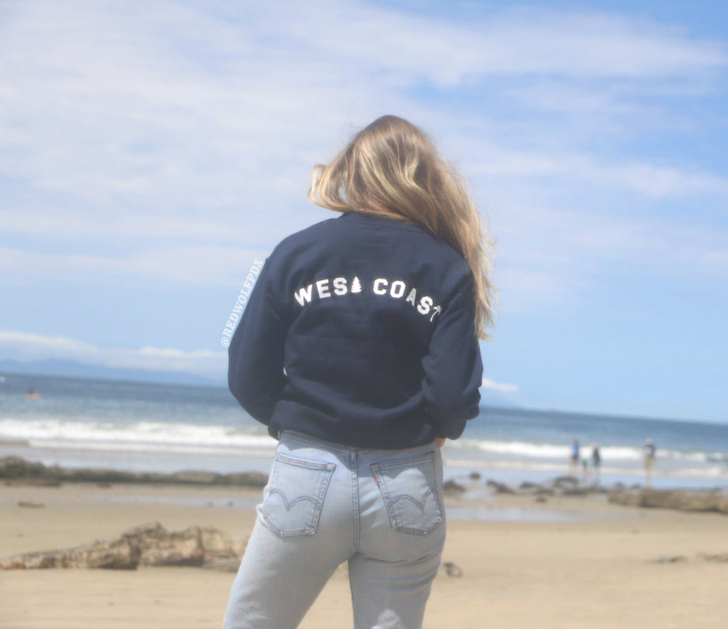 navy blue crewneck with text 'WEST COAST' text across the back being worn on sunny coastline