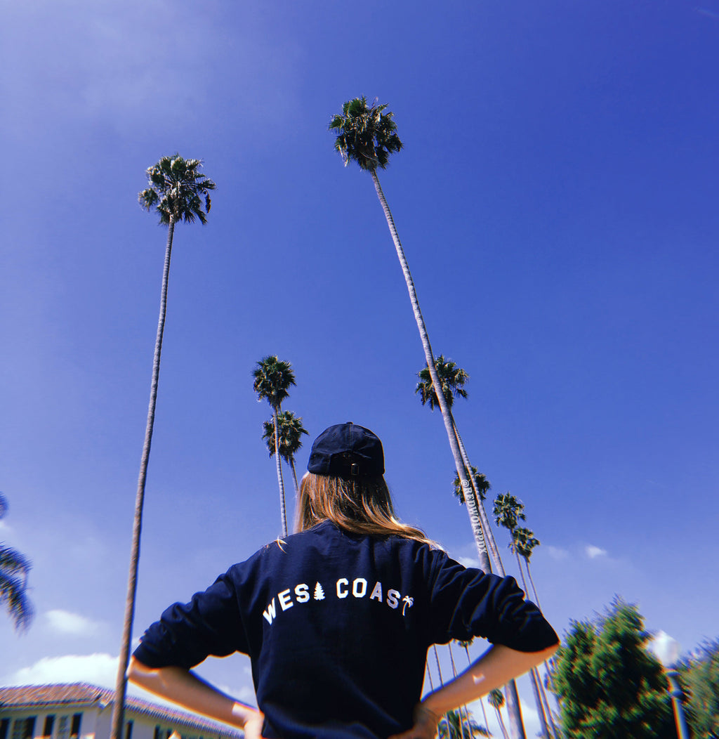 navy blue crewneck with text 'WEST COAST' text across the back being worn in sunny landscape with palm trees