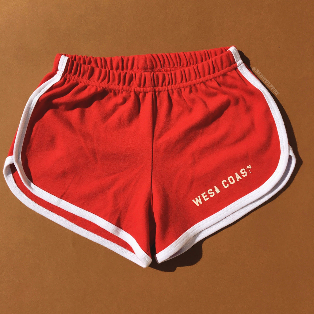 red track shorts with white trim with text 'WEST COAST' in white ink screen printed along edge of leg on a brown background