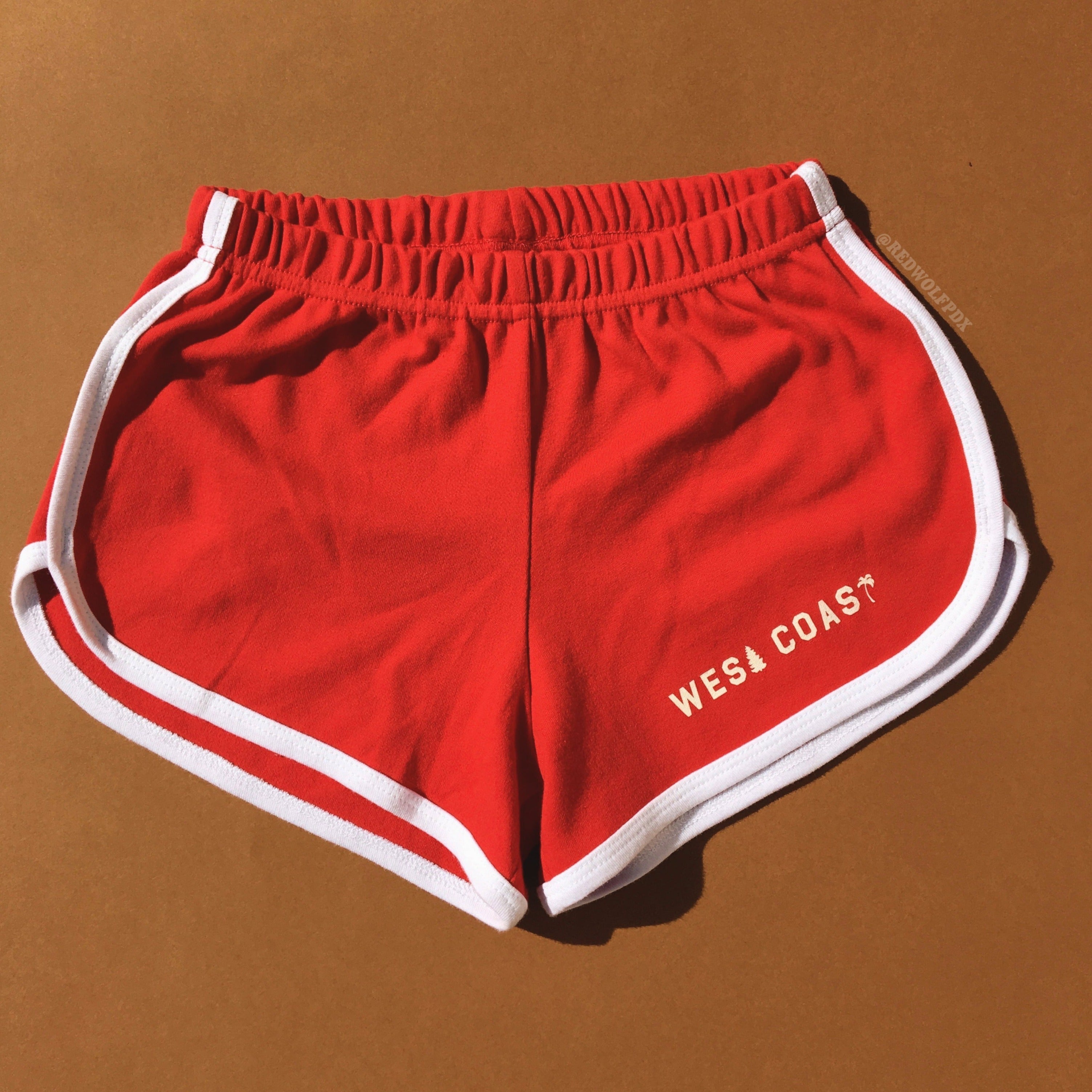 red track shorts with white trim with text 'WEST COAST' in white ink screen printed along edge of leg on a brown background