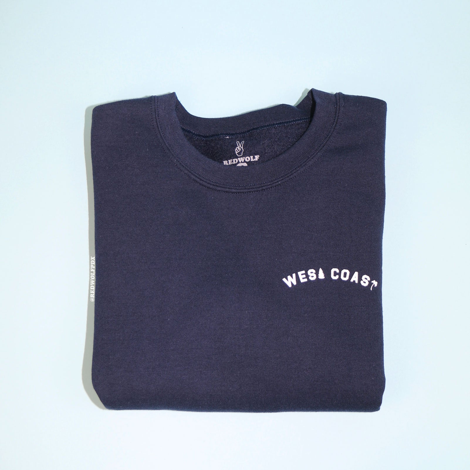 Folded navy blue crewneck sweatshirt with the text 'WEST COAST' printed in white.
