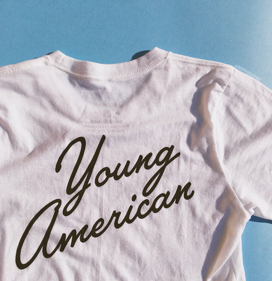 white graphic tee with text 'Young American' in a vintage inspired font screen printed across the back of tee against a blue backdrop