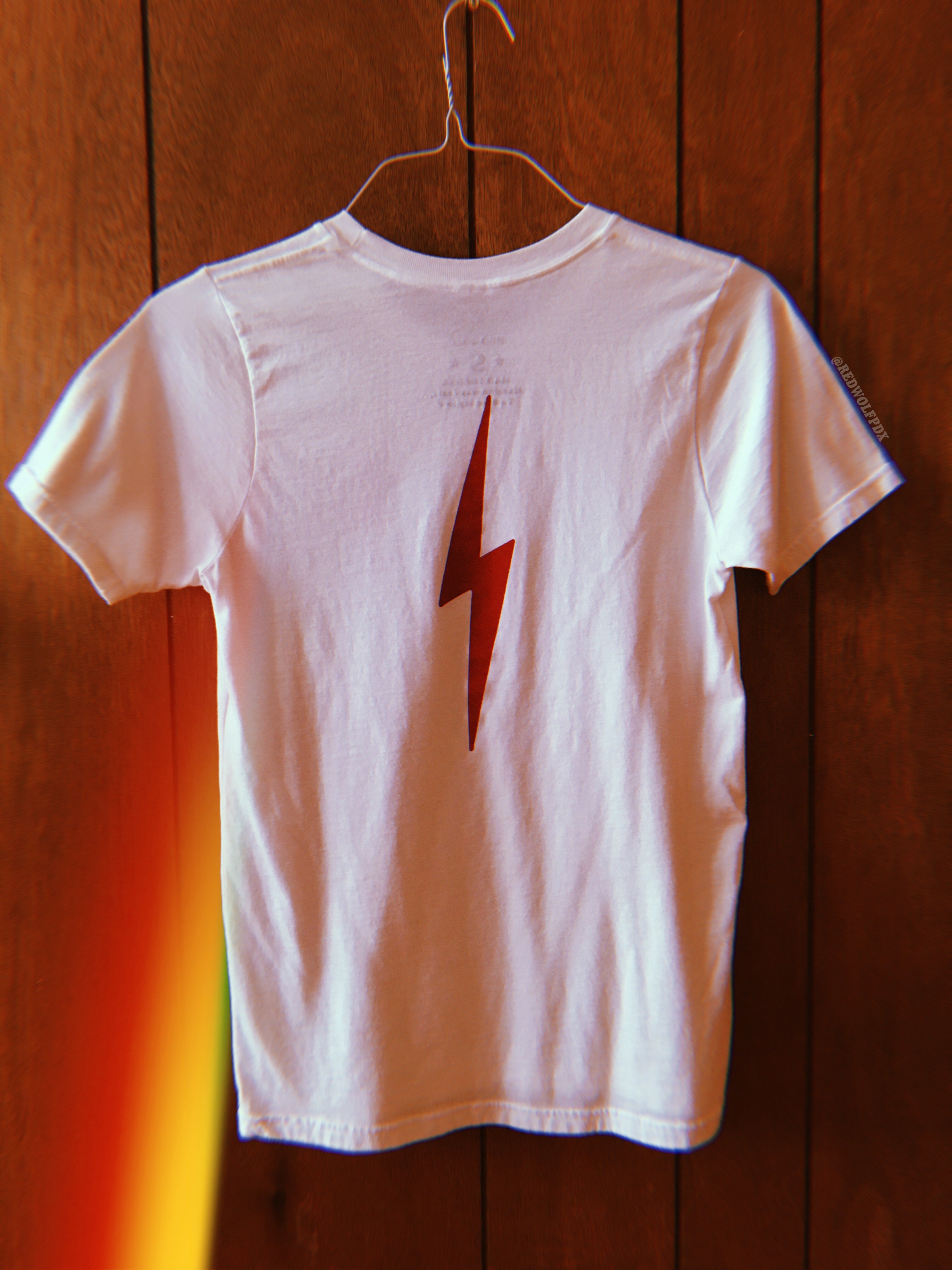 White graphic tee with lightening bolt in vintage red ink screen printed down the back of tee hanging from a hanger on a 70s wood panel wall