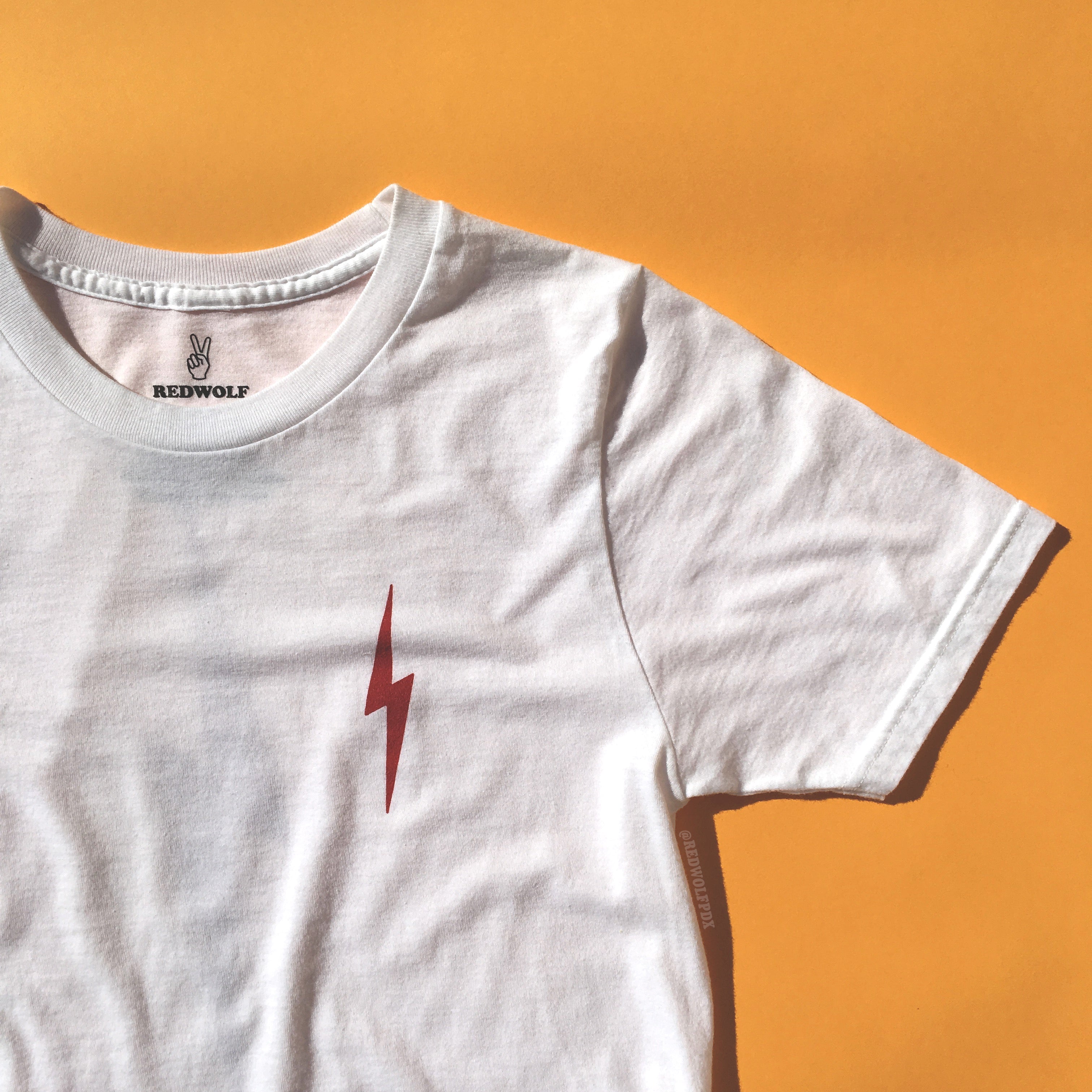 White graphic tee with lightening bolt in vintage red ink screen on front corner of chest against a vintage yellow background